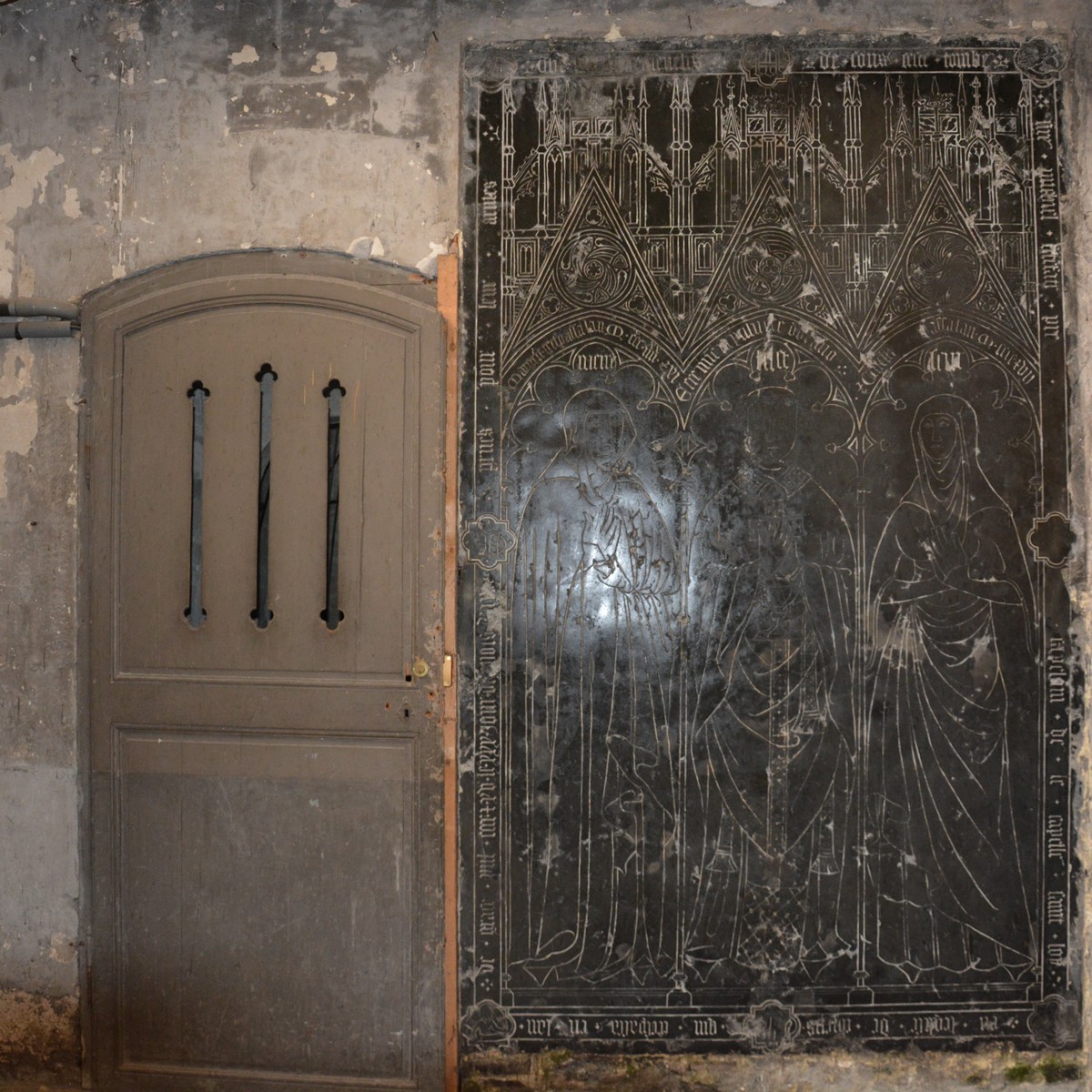  encastrée dans le mur du fond de la nef, une grande pierre bleue apparaît aux regards contenant trois personnages, abrités par de riches arcades à engrélures, couronnées de gables aigus à rosaces, de clochetons et de contreforts 
dont l'ensemble forme une gracieuse  architecture.
Le personnage du milieu est le chapelain Mchel Ficefate, décédé en 1415, et qui obtint la permission d'enterrer dans sa propre tombe sa mère et sa sœur, figurées à droite et à gauche et décédées avant lui, en 1391 et 1407.
Au pied de cette dalle et dans le pavé, une dernière pierre tombale dont l'inscription est fruste représente deux prêtres dont la tête et les mains sont de marbre blanc.