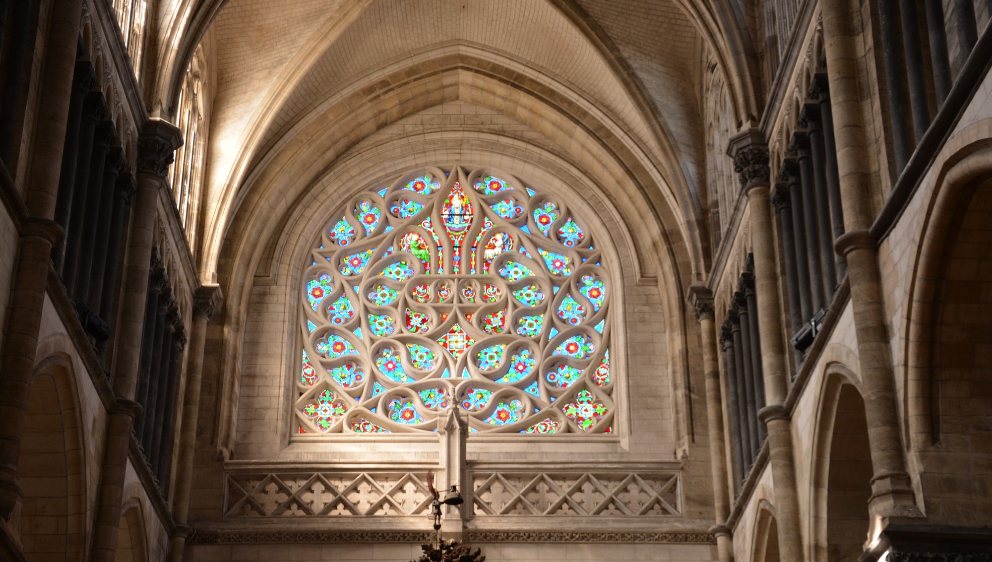 Cathédrale de Saint-Omer | rosace Nord | Rose à remplage supérieur en forme de fleur de lys. Deux écoinçons dans le bas forment une base horizontale. Verre à dominante rouge. Tout autour, multiples mouchettes entremêlées, à dominante bleue. Quadrilobes aux angles inférieurs sur pointe carrée.