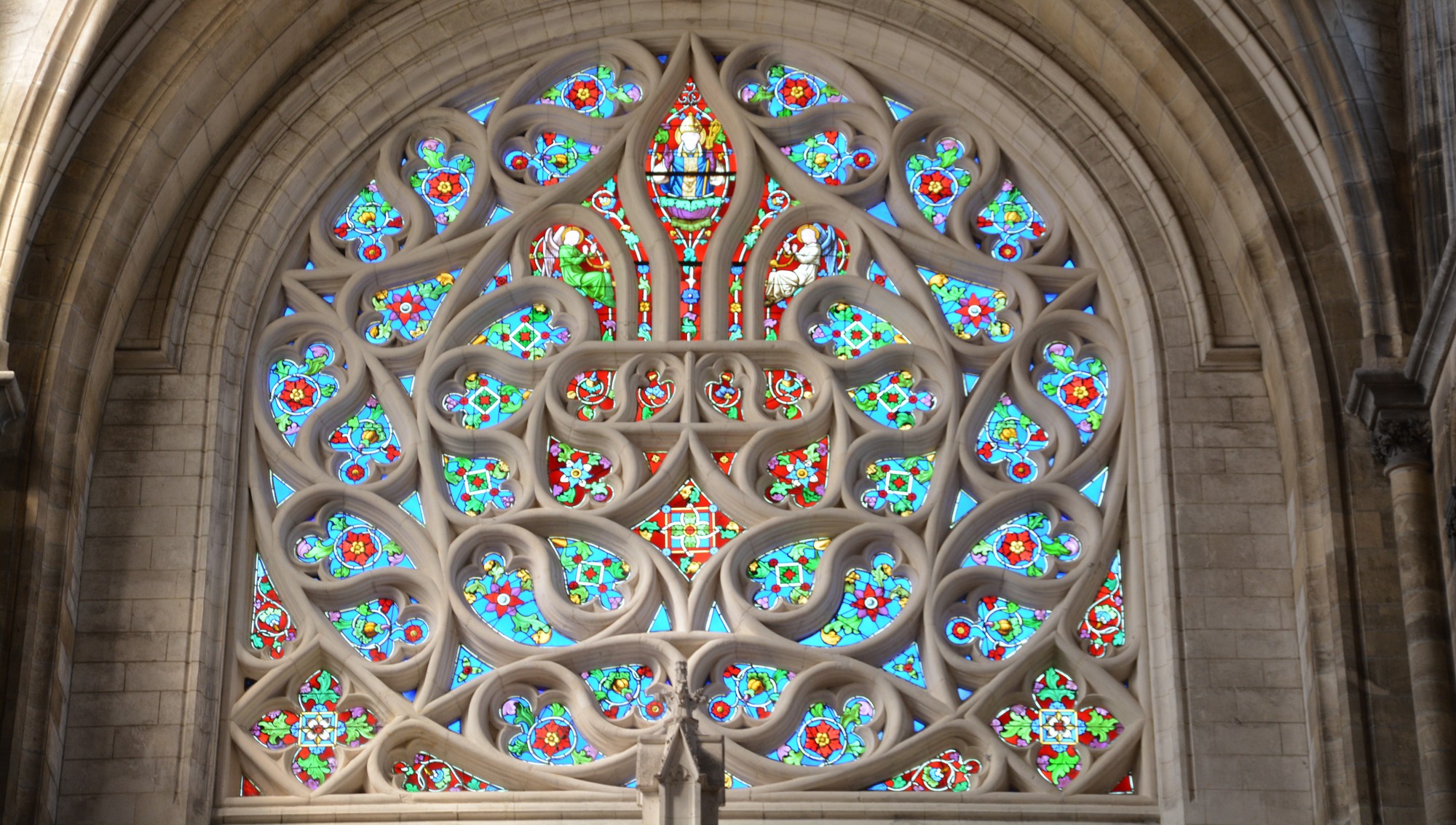 Cathédrale de Saint-Omer | rosace Nord | Rose à remplage supérieur en forme de fleur de lys. Deux écoinçons dans le bas forment une base horizontale. Verre à dominante rouge. Tout autour, multiples mouchettes entremêlées, à dominante bleue. Quadrilobes aux angles inférieurs sur pointe carrée.